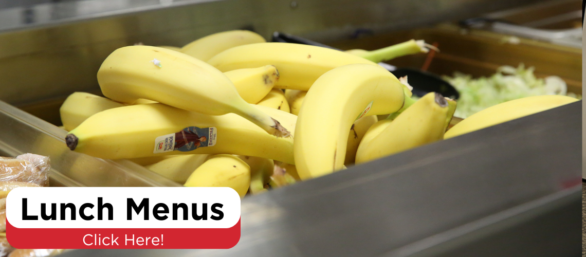 A close-up view of a bunch of ripe bananas placed in a metallic container, likely in a cafeteria or a similar setting.