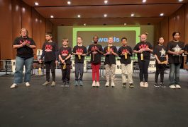Group of young people standing on stage in front of microphones, smiling at camera, wearing black shirts with red and white