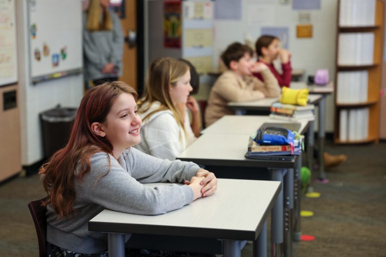 Students seated at desks in a classroom, looking towards the camera with smiles on their faces.