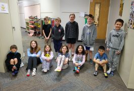 Students seated at desks in a classroom, looking towards the camera with smiles on their faces.