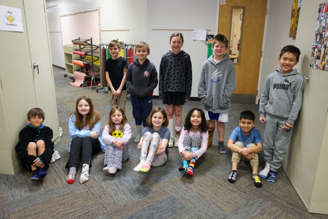Students seated at desks in a classroom, looking towards the camera with smiles on their faces.