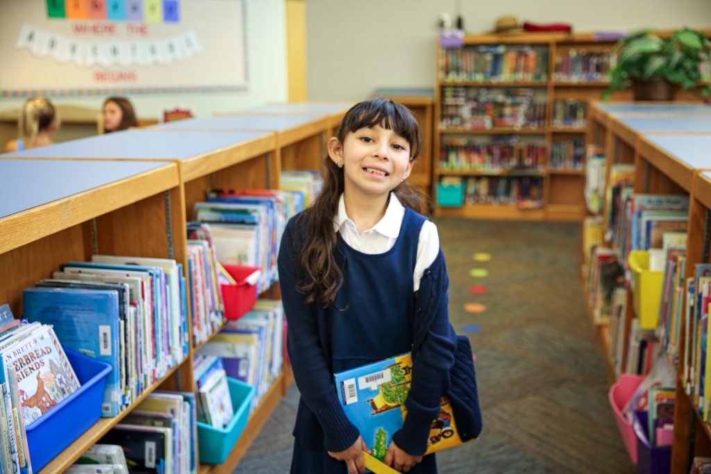 Young girl in a library, standing in front of bookshelves filled with books. She is wearing a blue sweater and holding a