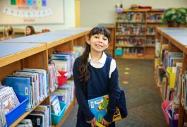 Young girl in a library, standing in front of bookshelves filled with books. She is wearing a blue sweater and holding a